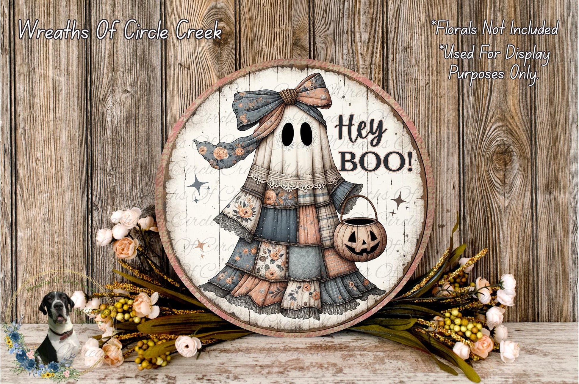 a Halloween-themed decoration with a ghost wearing a dress and holding a pumpkin. The ghost has a bow on its head and the text "Hey Boo!" written on it. The decoration is displayed on a wooden surface, surrounded by flowers and greenery.