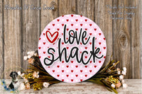 a pink circular sign with the words "Love Shack" written on it, surrounded by a floral arrangement and a wooden background.