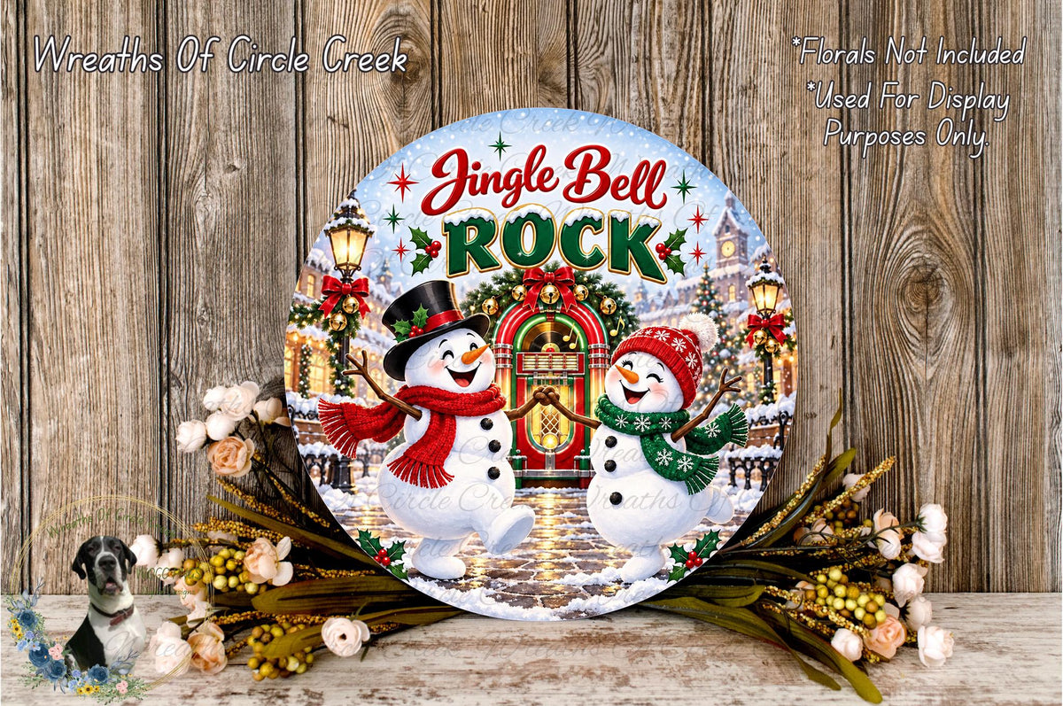 A round decorative plate featuring a winter scene with two snowmen, a gingerbread house, and Christmas lights. The plate has the text "Jingle Bell Rock" and is surrounded by flowers and greenery.