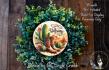 a wreath with a welcome sign, surrounded by various succulents and cacti, including a pair of cowboy boots. The wreath is displayed on a wooden surface, and there is a small dog in the bottom right corner.