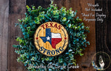 A wreath made of green foliage and a wooden sign with the text "Texas Strong" in the center, surrounded by a wooden background.