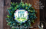 A wreath made of green leaves and a circular sign with the text "Living My Best Life by Par" is displayed on a wooden surface.
