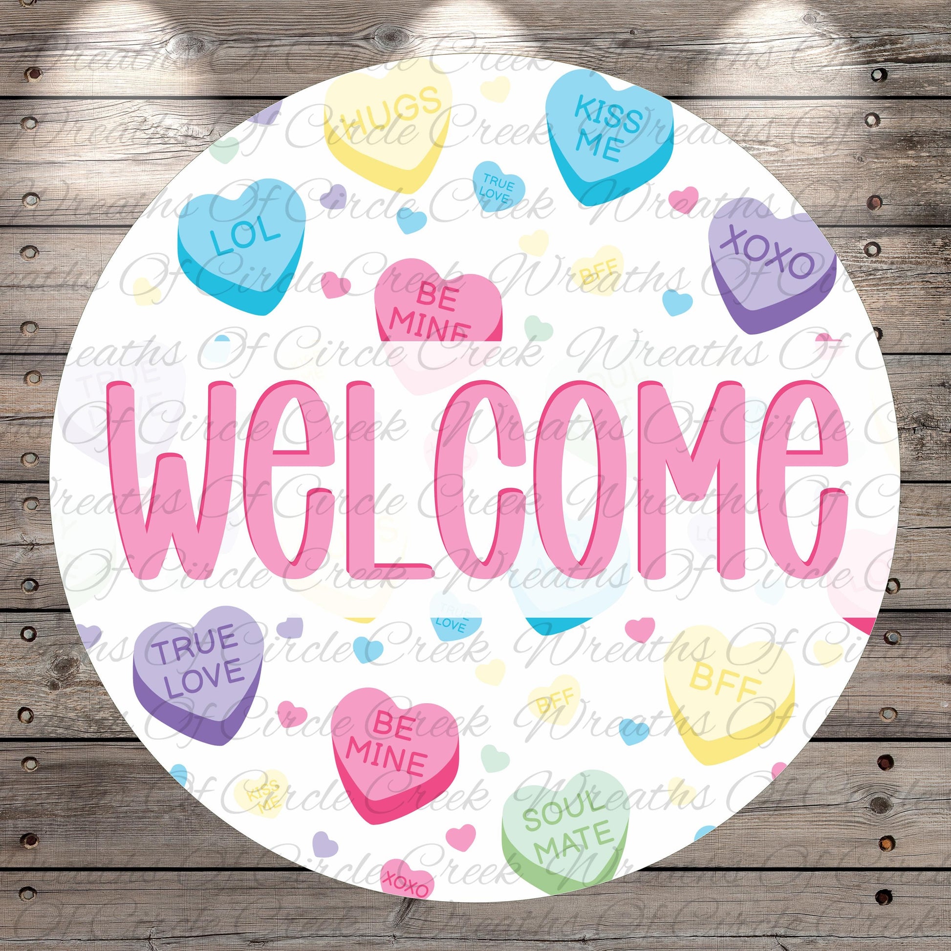 The image is a circular welcome sign with the word "WELCOME" written in large pink letters. The sign is surrounded by various heart-shaped candies with different messages such as "LOL", "KISS ME", "HUGS", "TRUE LOVE", "BE MINE", "SOUL MATE", and "KINGS". The background of the sign is a wooden surface with a repeating pattern of these heart-shaped candies.