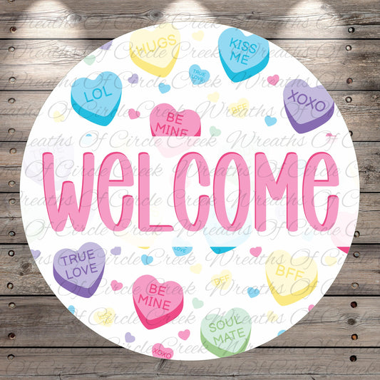 The image is a circular welcome sign with the word "WELCOME" written in large pink letters. The sign is surrounded by various heart-shaped candies with different messages such as "LOL", "KISS ME", "HUGS", "TRUE LOVE", "BE MINE", "SOUL MATE", and "KINGS". The background of the sign is a wooden surface with a repeating pattern of these heart-shaped candies.