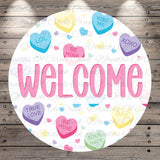 The image is a circular welcome sign with the word "WELCOME" written in large pink letters. The sign is surrounded by various heart-shaped candies with different messages such as "LOL", "KISS ME", "HUGS", "TRUE LOVE", "BE MINE", "SOUL MATE", and "KINGS". The background of the sign is a wooden surface with a repeating pattern of these heart-shaped candies.