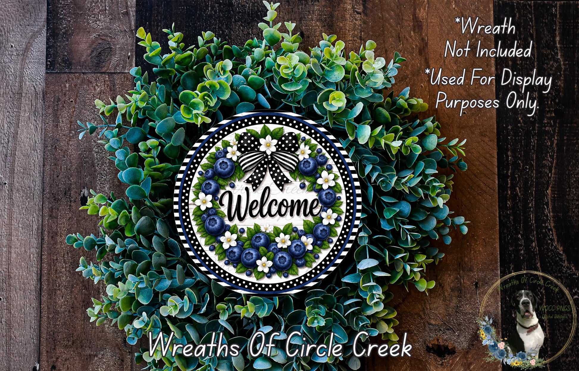 a wreath made of various green and blue foliage, with a white and blue sign that says "Welcome" in the center. The wreath is displayed on a wooden surface, and there is a small dog in the bottom right corner of the image.