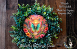 A wreath made of green leaves and flowers, with a colorful butterfly design in the center. The wreath is displayed on a wooden surface, and there is a small dog in the bottom right corner.