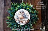 A wreath featuring a painted owl sitting on a log, surrounded by green foliage.