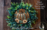 A wreath made of various green leaves and a leopard print pumpkin, with a dog in the background.