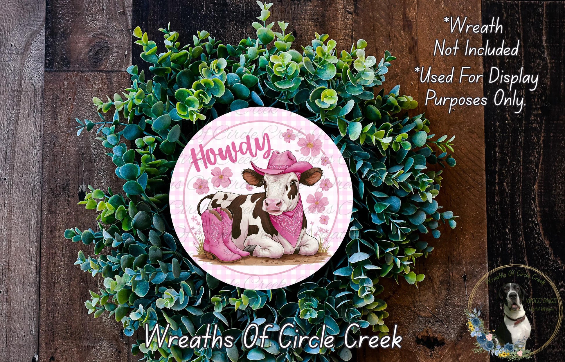 a wreath with a cow wearing a pink hat and scarf, surrounded by green foliage. The wreath is displayed on a wooden surface, and there is a small dog in the bottom right corner of the image.