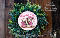 a wreath with a cow wearing a pink hat and scarf, surrounded by green foliage. The wreath is displayed on a wooden surface, and there is a small dog in the bottom right corner of the image.