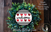 a wreath made of green leaves and red hearts, with the word "welcome" written on it. The wreath is displayed on a wooden surface, and there is a small dog in the bottom right corner of the image.