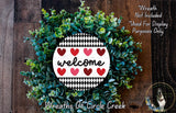 a wreath made of green leaves and red hearts, with the word "welcome" written on it. The wreath is displayed on a wooden surface, and there is a small dog in the bottom right corner of the image.
