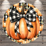 A large, orange pumpkin with a black and white checkered bow tied around it, surrounded by additional orange pumpkins and leaves against a wooden background.