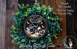 A wreath made of green leaves and a black cat wearing a witch's hat, with a clock face in the center.
