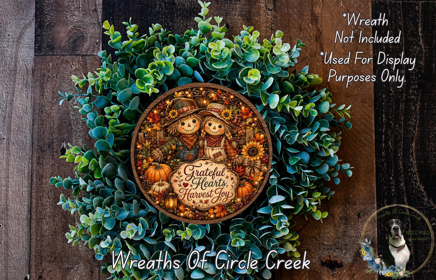 a wreath with a central design of two scarecrows and a pumpkin, surrounded by a lush green wreath. The wreath is displayed on a wooden surface, and there is a small dog in the bottom right corner of the image.
