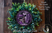 A wreath made of green leaves and purple flowers, with a witch's hat and boots on top, and a purple and gold book on a shelf.