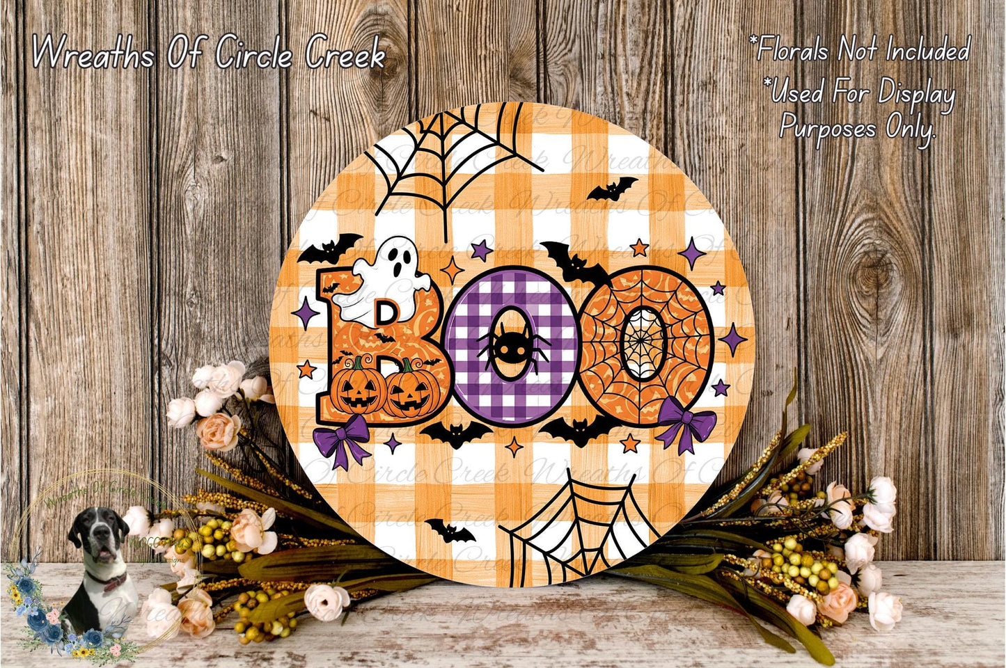 a Halloween-themed decoration with a large circular sign that says "Boo" in orange and purple colors, surrounded by a spider web, bats, and pumpkins. The decoration is placed on a wooden surface, with a wreath of flowers and a spooky spider decoration nearby.