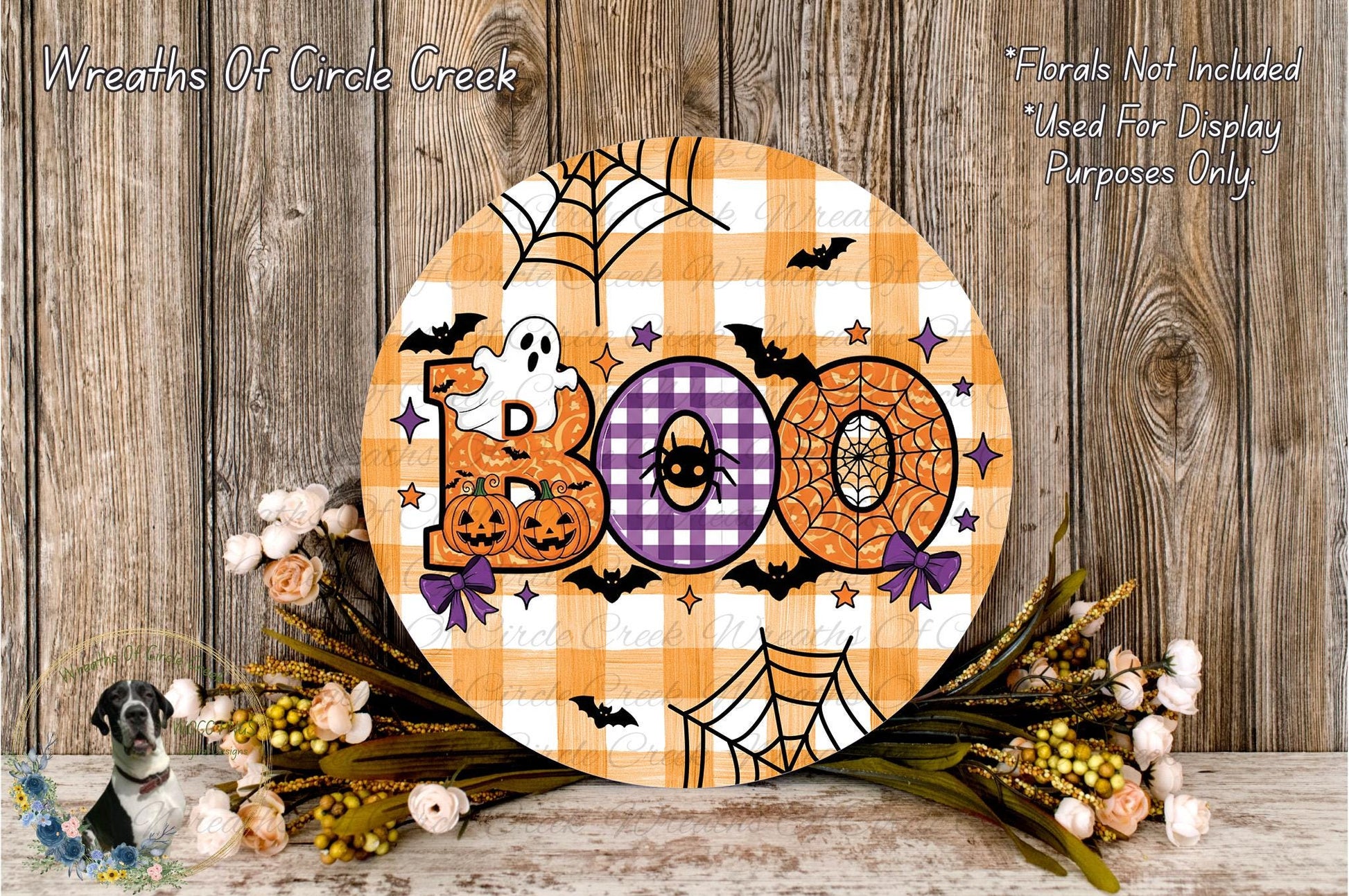 a Halloween-themed decoration with a large circular sign that says "Boo" in orange and purple colors, surrounded by a spider web, bats, and pumpkins. The decoration is placed on a wooden surface, with a wreath of flowers and a spooky spider decoration nearby.