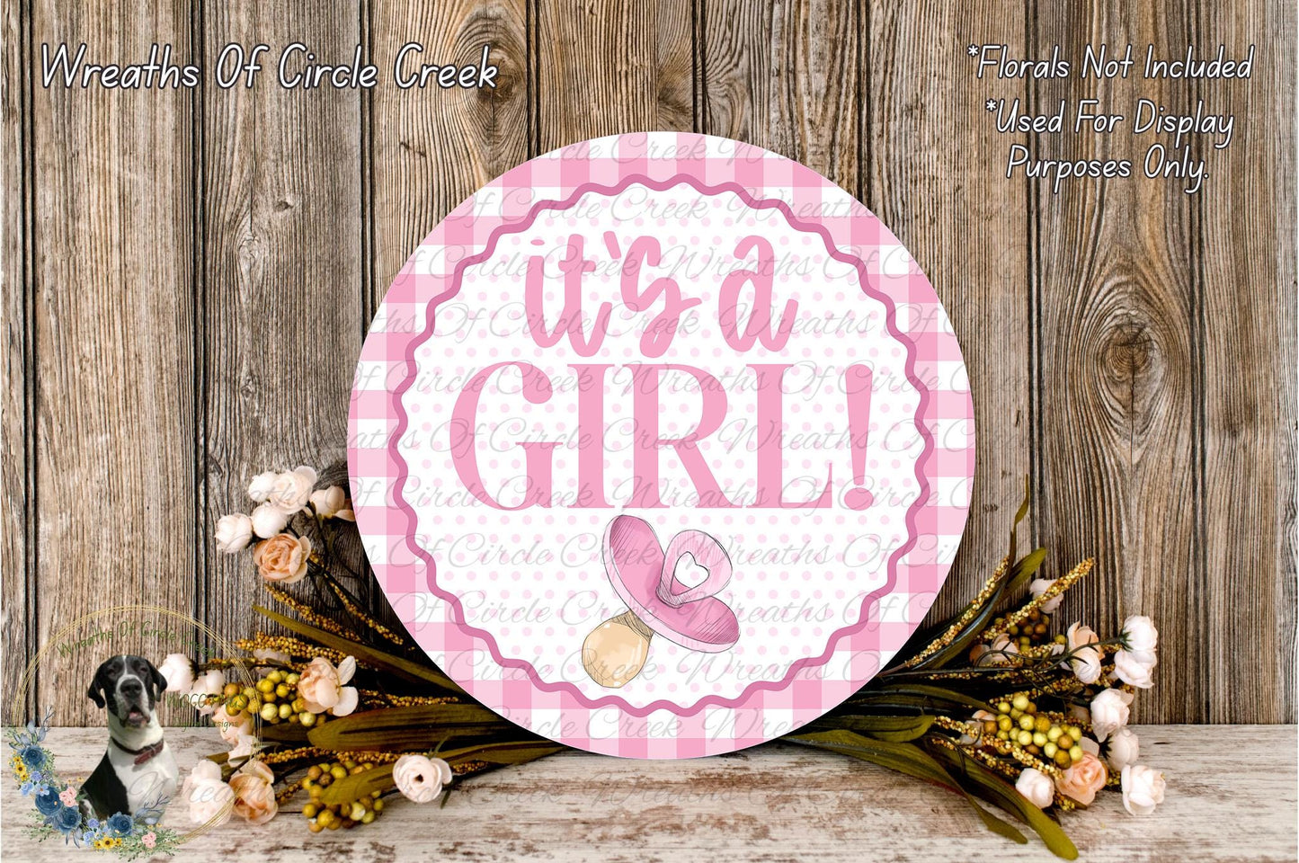 a round sign with the text "It's a girl!" surrounded by a floral arrangement and a wooden background.