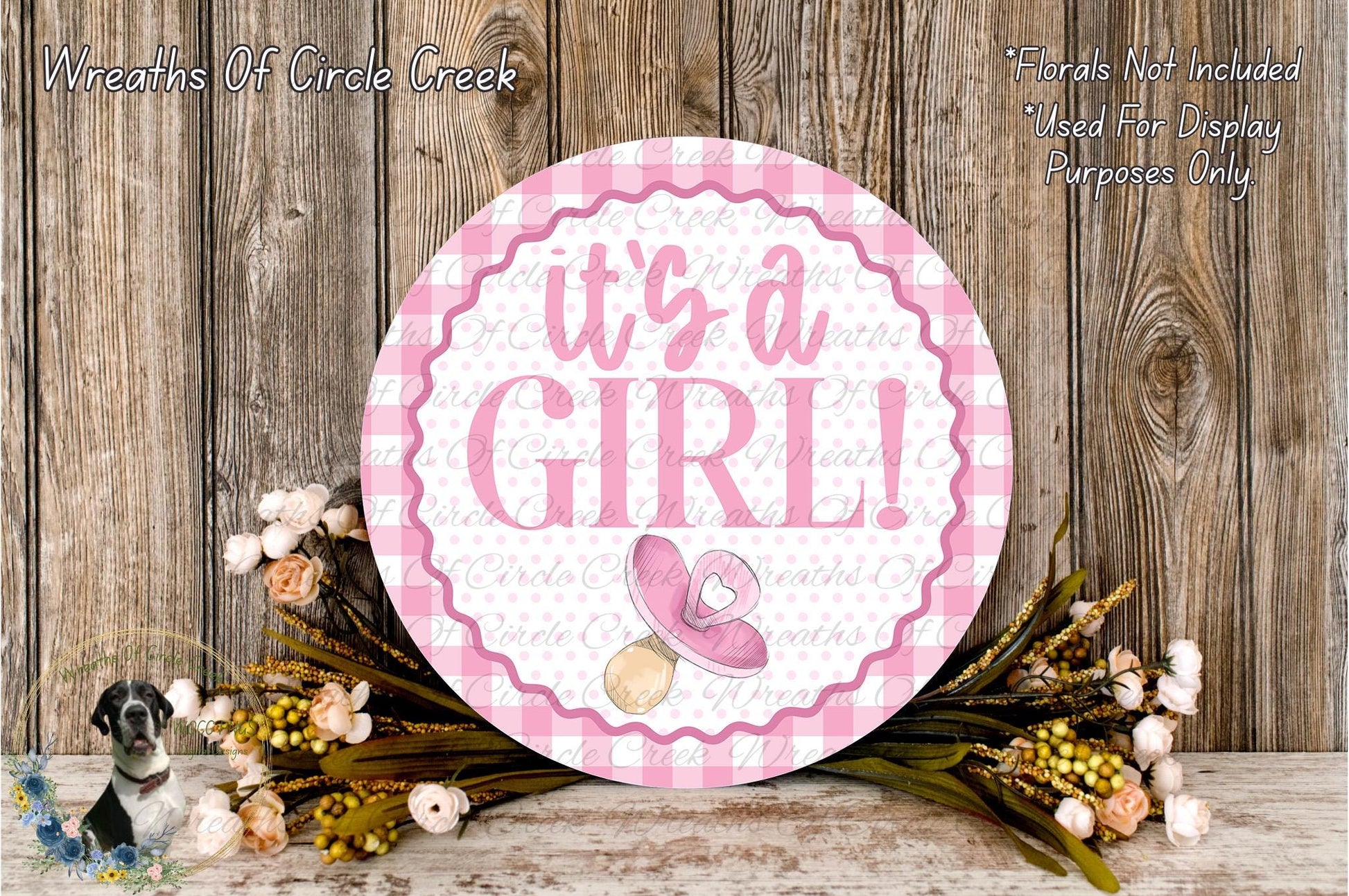 a round sign with the text "It's a girl!" surrounded by a floral arrangement and a wooden background.