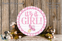 a round sign with the text "It's a girl!" surrounded by a floral arrangement and a wooden background.