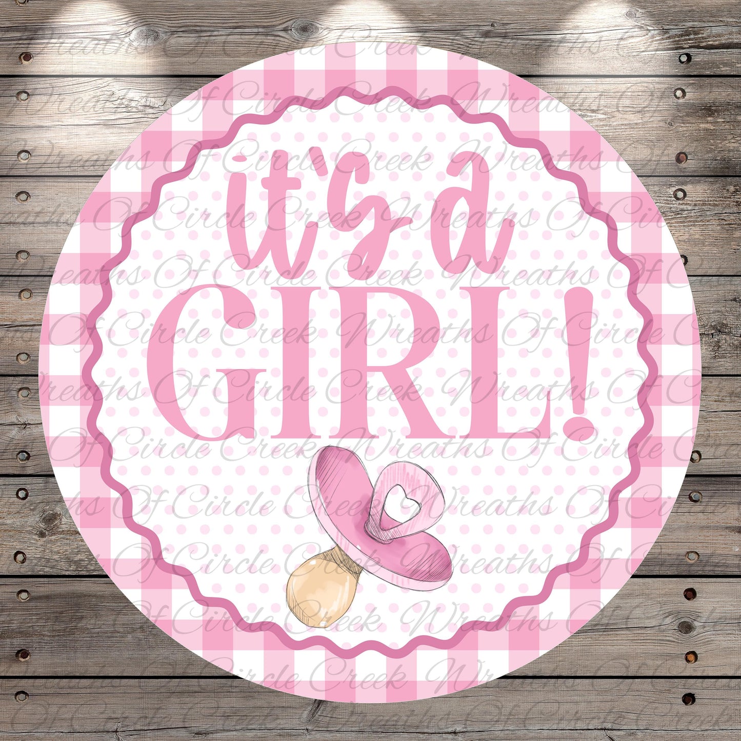 A pink and white circular sign with the text "It's a girl!" and a pacifier icon, placed on a wooden background.