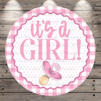 A pink and white circular sign with the text "It's a girl!" and a pacifier icon, placed on a wooden background.