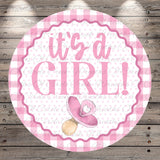 A pink and white circular sign with the text "It's a girl!" and a pacifier icon, placed on a wooden background.