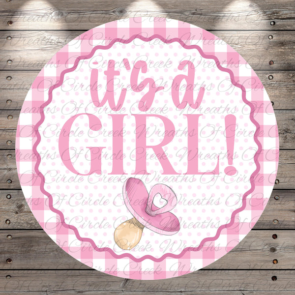 A pink and white circular sign with the text "It's a girl!" and a pacifier icon, placed on a wooden background.