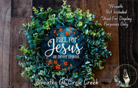 a wreath made of green leaves and a blue circular sign with the text "Fall for Jesus he never leaves" against a wooden background.