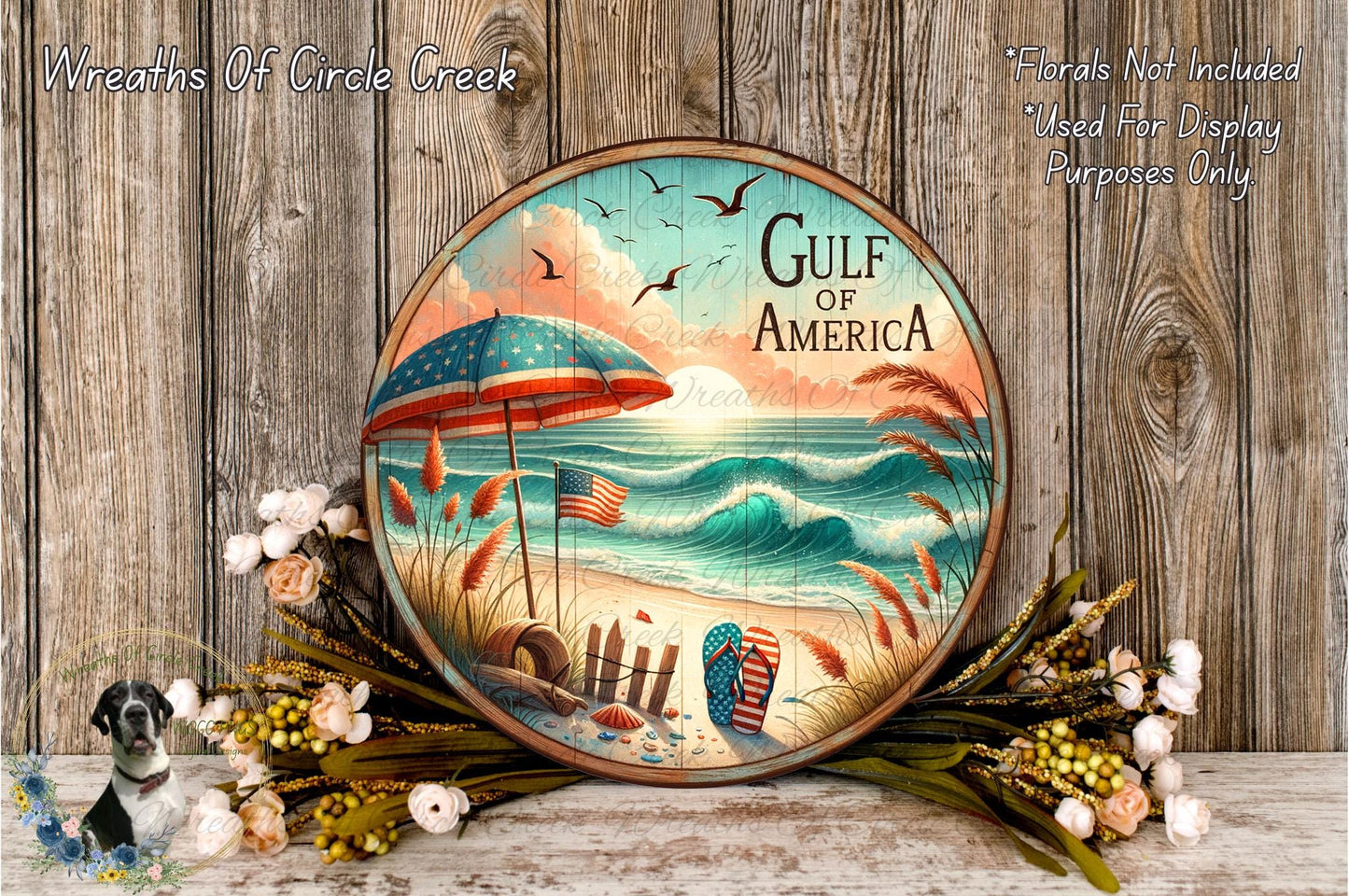 A round wooden plaque with a beach scene and an American flag, surrounded by flowers and placed on a wooden surface.