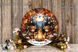 A circular wall hanging depicting two cartoon cows sitting around a campfire, with a full moon and autumn leaves in the background. The cows are wrapped in blankets and appear to be enjoying the warmth of the fire. The wall hanging is surrounded by flowers and greenery, creating a cozy and festive atmosphere.