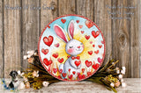 A round, decorative wreath with a white rabbit holding red hearts, surrounded by flowers and greenery, placed on a wooden surface.