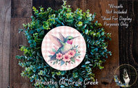 a wreath with a hummingbird and flowers, surrounded by greenery. The wreath is placed on a wooden surface.