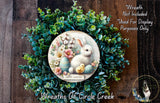 a wreath with a white rabbit and Easter eggs, surrounded by a lush green wreath. The wreath is displayed on a wooden surface, and there is a small dog in the bottom right corner of the image.