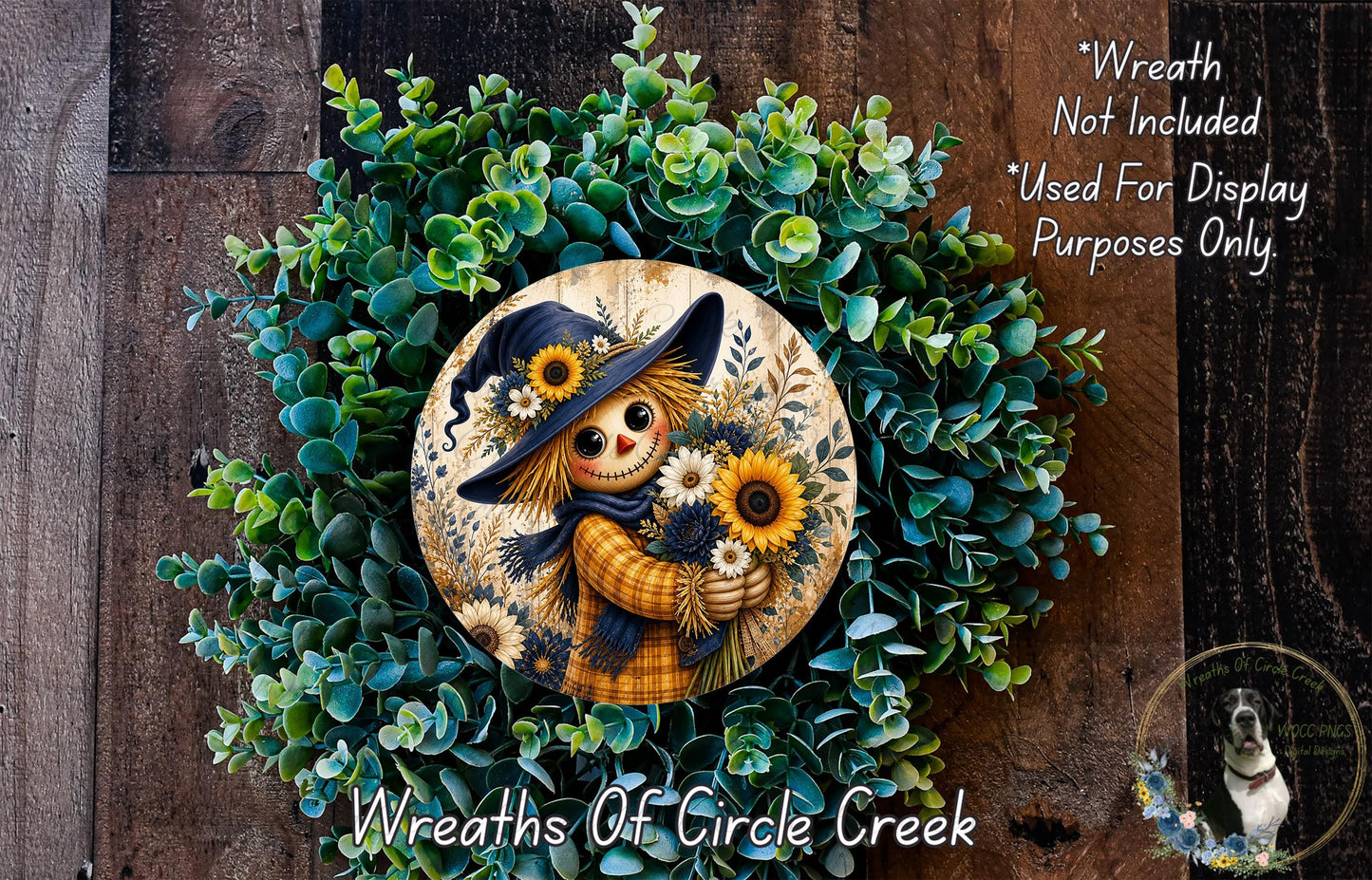 a wreath with a scarecrow figure holding flowers, surrounded by a lush green wreath. The wreath is displayed on a wooden surface, and there is a small dog in the bottom right corner of the image.