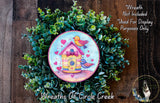 A wreath made of green leaves and flowers, featuring a small birdhouse painted on it.