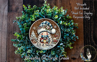 A wreath made of green foliage and featuring a smiling gnome wearing a blue and white hat, with the text "Let it snow" and "Wreaths of Circle Creek" displayed.