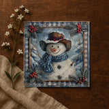 A framed painting of a smiling snowman wearing a hat and scarf, surrounded by winter elements like pine branches and berries, is displayed on a wooden surface.