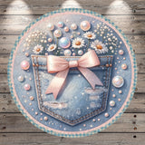 A round, decorative piece featuring a denim pocket with a pink bow, surrounded by white flowers and small bubbles, all set against a wooden background.