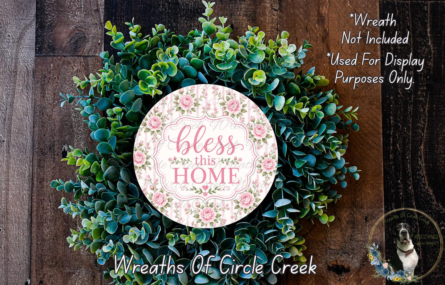 A wreath made of various green plants and flowers, with a pink circular sign that says "bless this home" in the center.