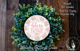 A wreath made of various green plants and flowers, with a pink circular sign that says "bless this home" in the center.