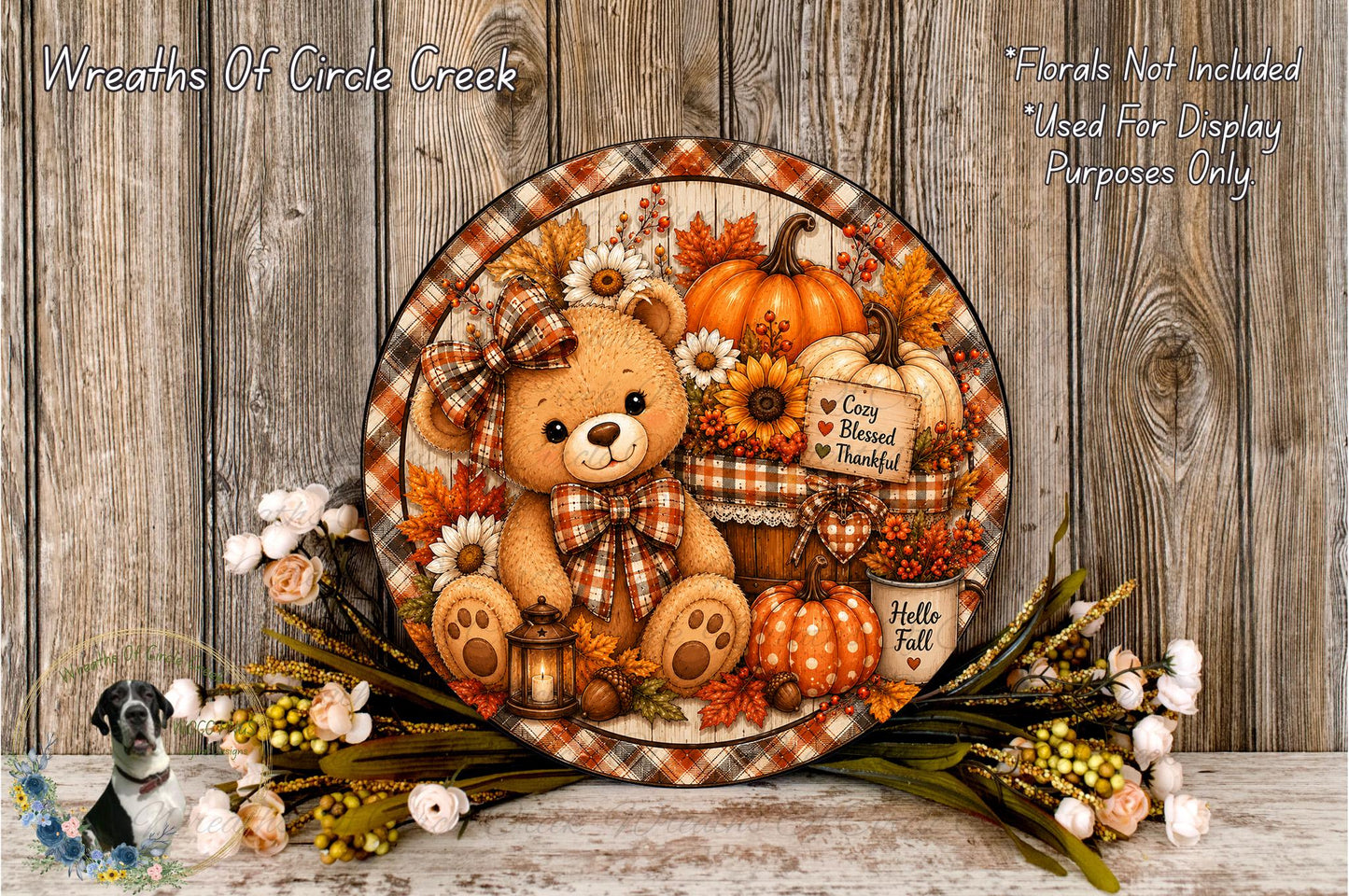 A decorative wreath featuring a teddy bear surrounded by autumn-themed elements such as pumpkins, flowers, and a lantern.