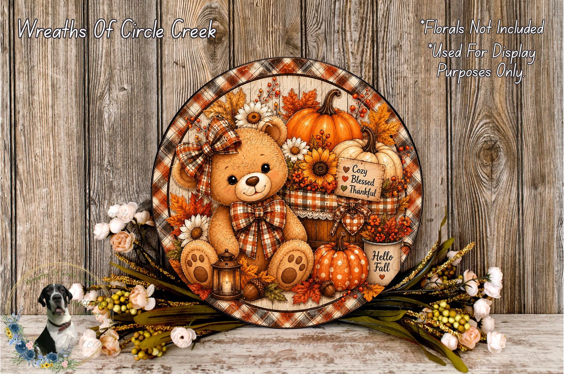 A decorative wreath featuring a teddy bear surrounded by autumn-themed elements such as pumpkins, flowers, and a lantern.