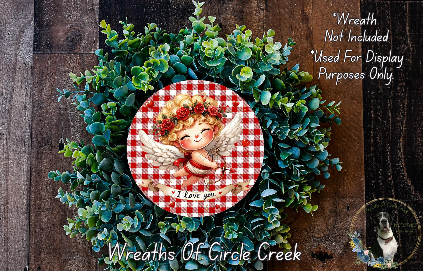 A wreath made of green leaves and flowers, with a red and white checkered border and a cherub figure in the center. The cherub is smiling and holding a heart.