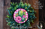 a wreath made of various green plants, including succulents and cacti, with a pink and white checkered border. The wreath is displayed on a wooden surface, and there is a small dog in the bottom right corner of the image.