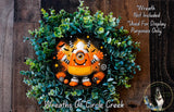a wreath with a large, colorful monster character at the center, surrounded by green foliage. The monster appears to be smiling and has a large eye on its face. The wreath is set against a wooden background, and there is a small dog visible in the bottom right corner of the image.