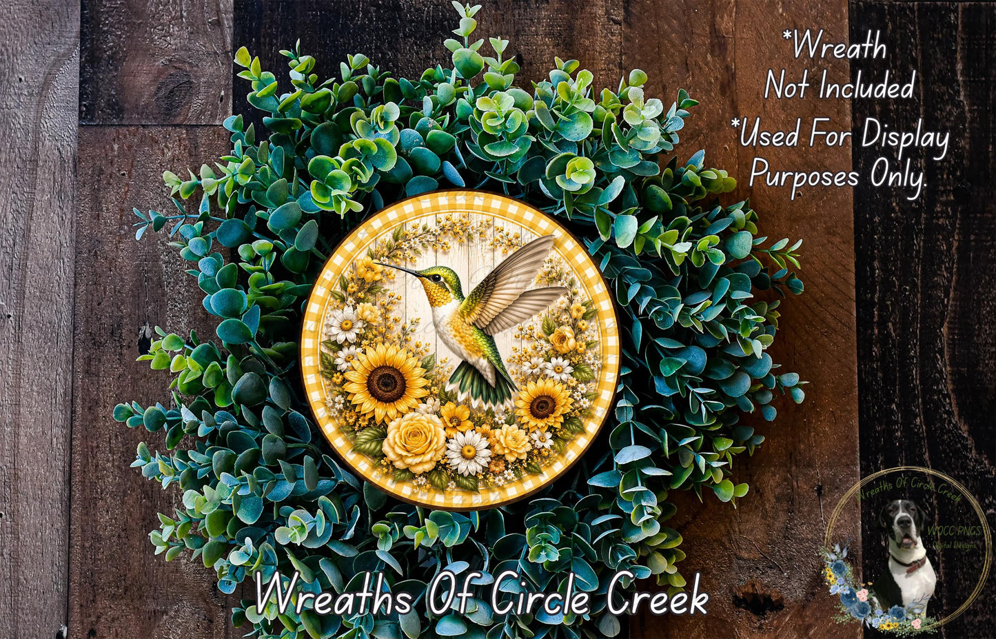 A decorative wreath made of green foliage and yellow flowers, featuring a hummingbird in flight. The wreath is displayed on a wooden surface, and there is a text overlay that reads "Wreath Not Included * Used For Display Purposes Only".