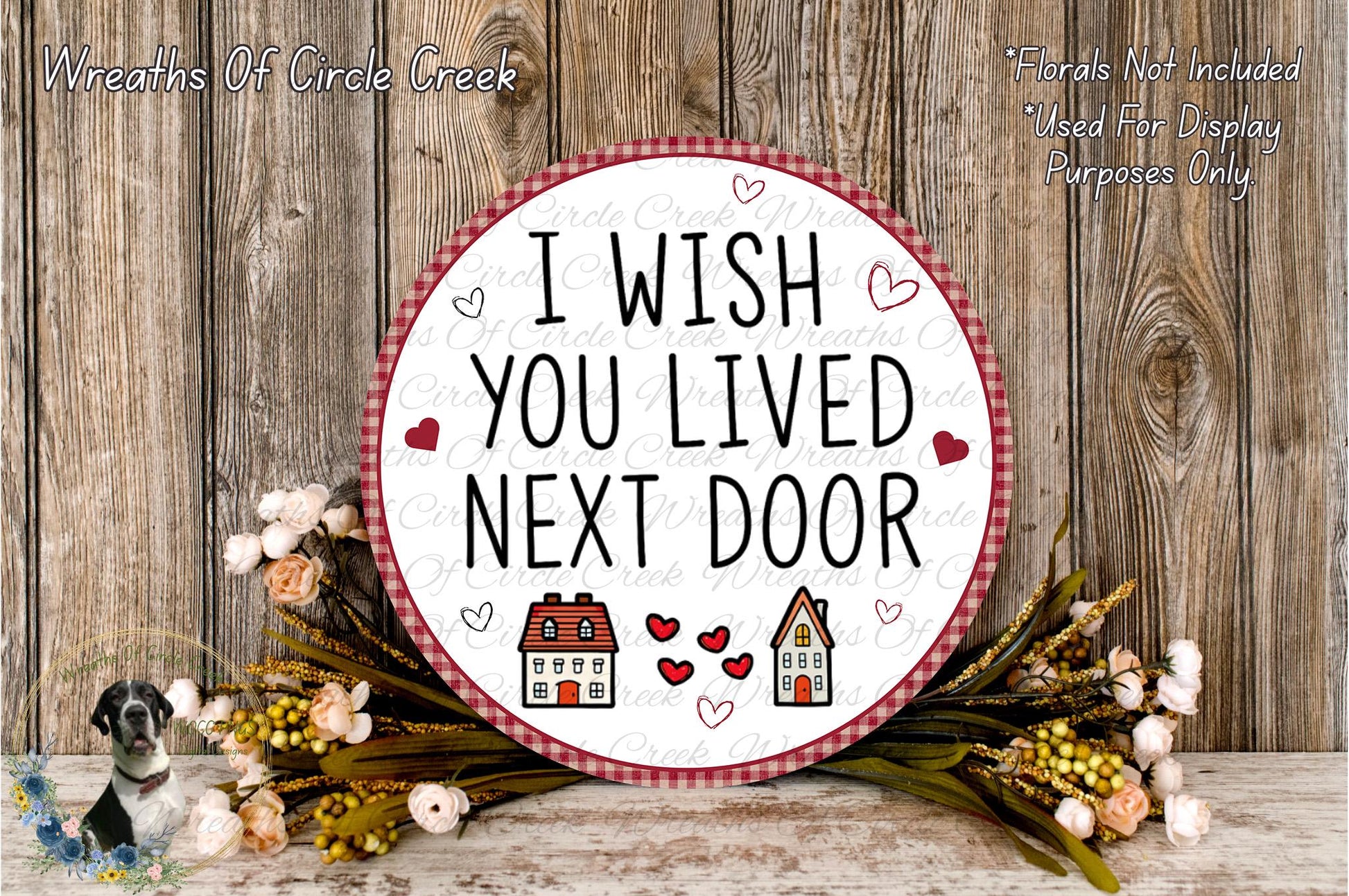 a round sign with the text "I WISH YOU LIVED NEXT DOOR" surrounded by a floral arrangement and a wooden background.